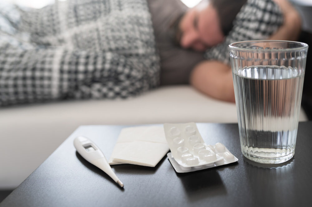 Meds, medical thermometer and glass of water on nightstand against person sleeping in bed