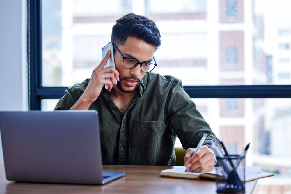 Shot of a young businessman writing notes while talking on a cellphone in an office