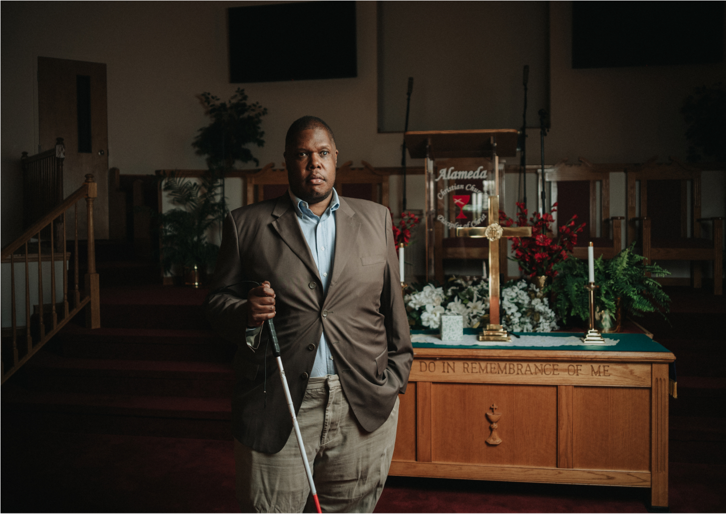 Jonathan standing in front of a church altar which is adorned in a cross, candles, and flowers, holding his white cane with one hand, and the other in his pocket. Looking at the camera.