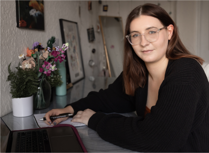 Simone sitting at her desk at home holding a pen next to her laptop and a vase of flowers, smiling slightly at the camera.