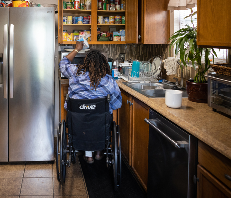 Melanie in her kitchen reaching into the top cabinet from her wheelchair