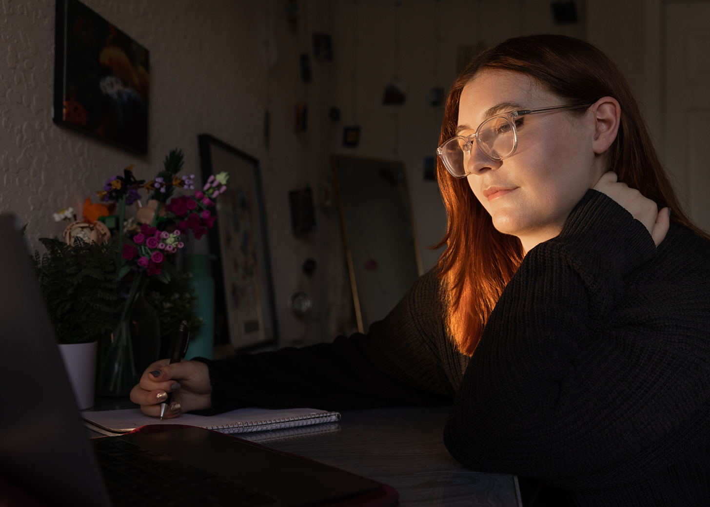 Simone sitting at her desk next to a vase of flowers, looking at the laptop and writing with a pen and notepad