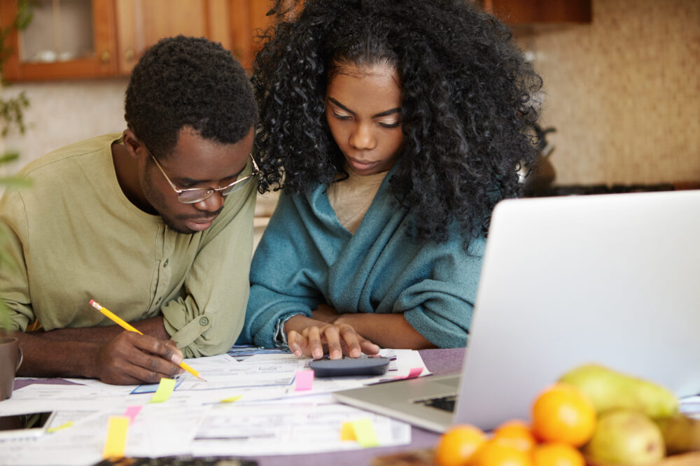 Unhappy African family facing financial stress, having big trouble while calculating domestic budget, frustrated with amount of expenses. Woman making calculations and her husband filling in papers
