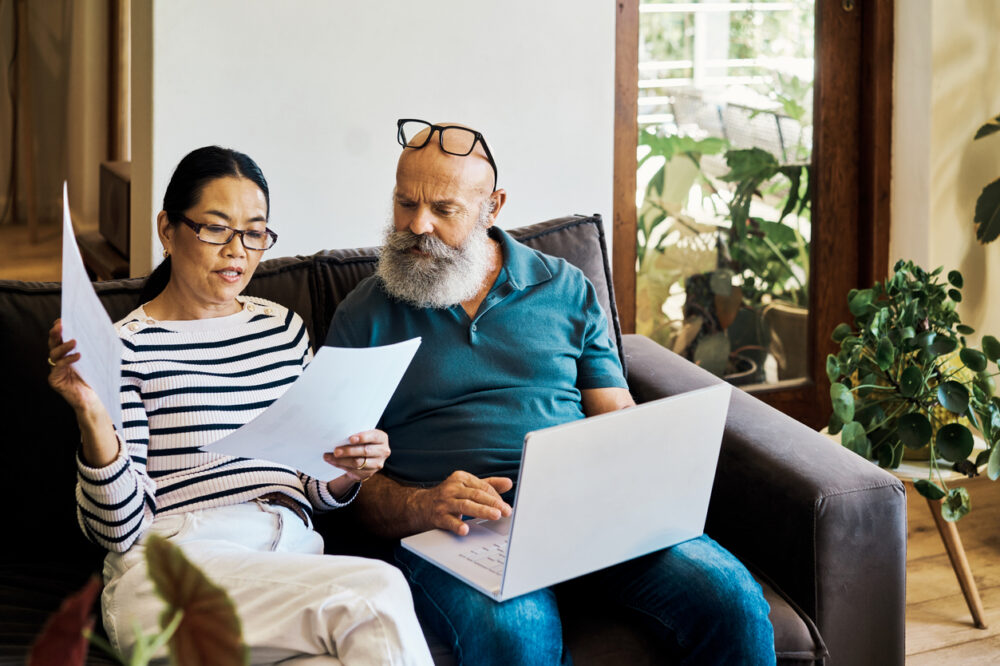 Senior interracial couple using a laptop and going retirement documents online on the couch at home.