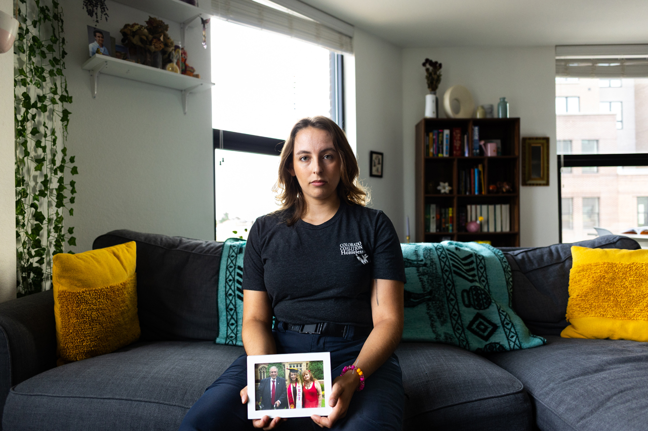 Angel sits on their couch holding a framed photo of them and their parents at graduation