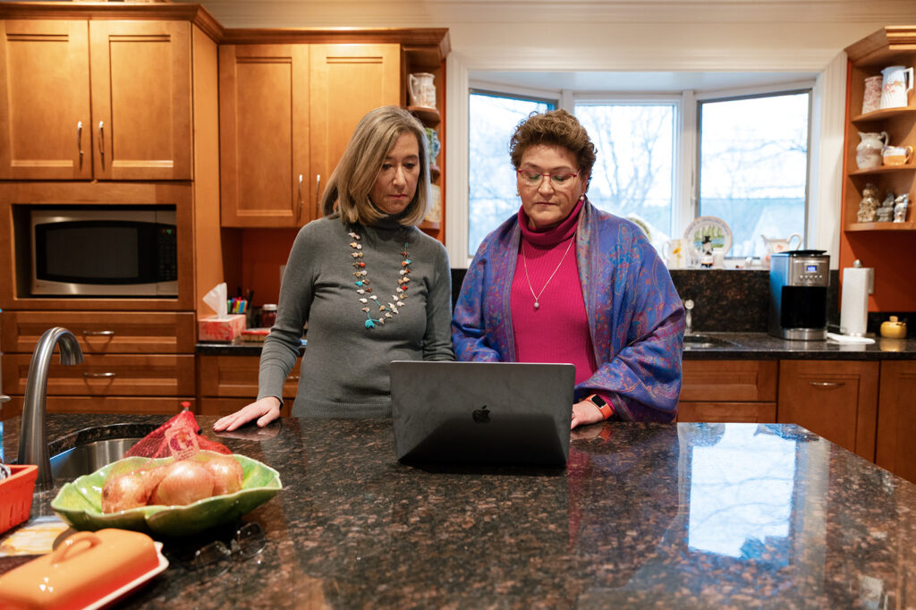 Elizabeth and Linda stand at the kitchen counter researching on the laptop.