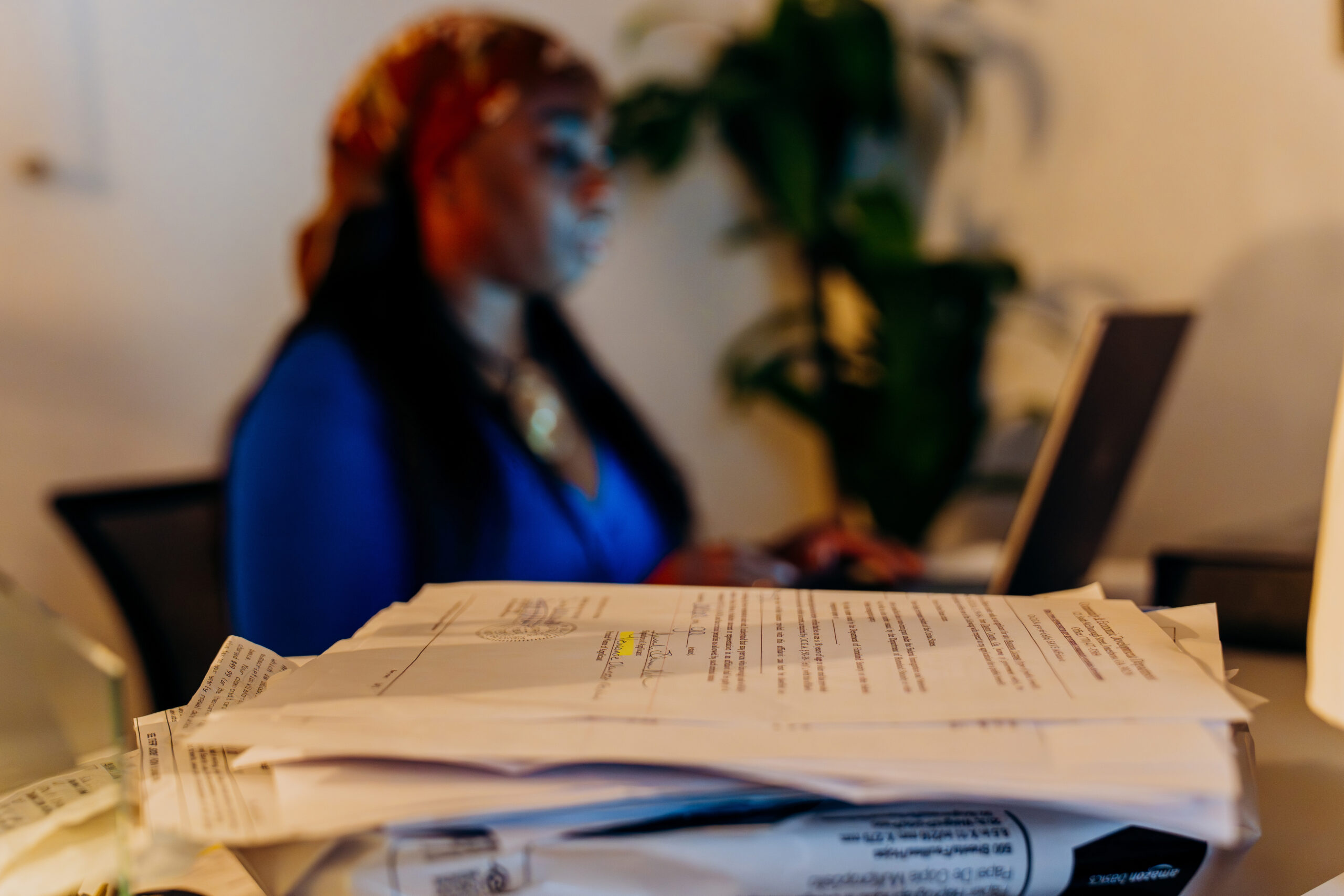 A stack of papers in the foreground with Wande in the background working on her laptop
