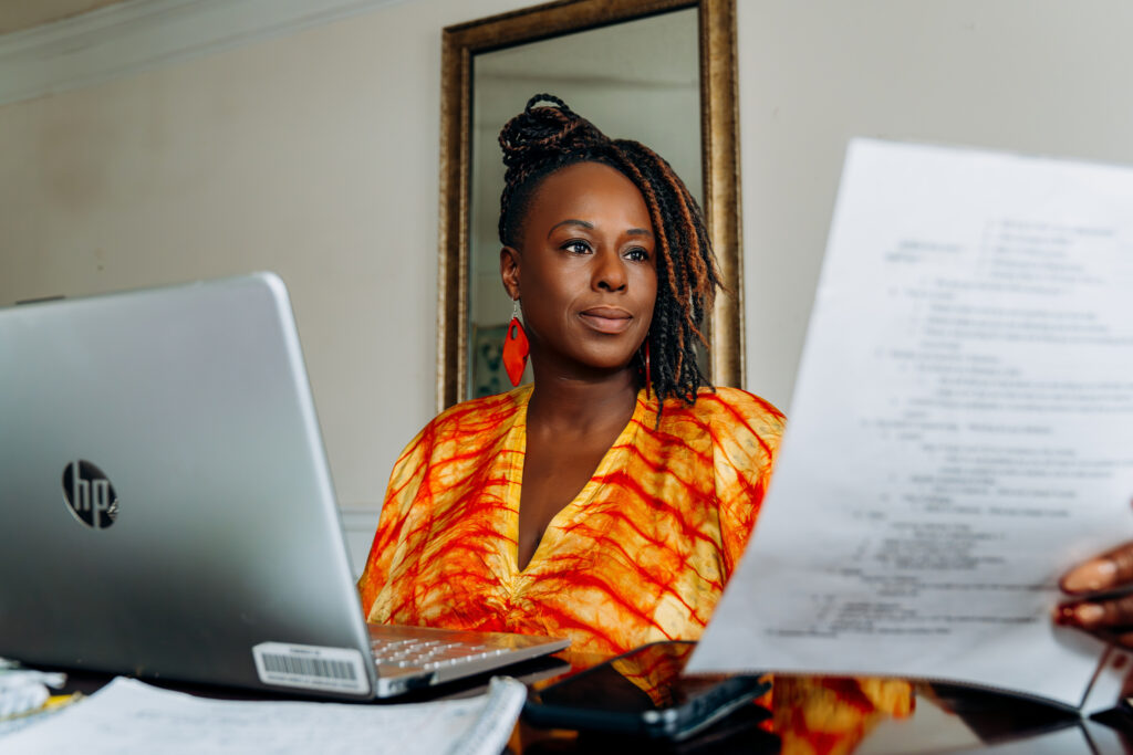 Ori sits at her home desk with an opened laptop and examines a paper in her hand