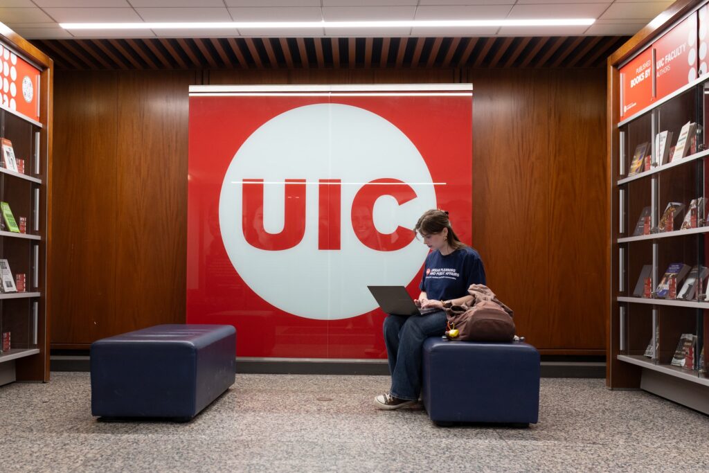 Margarita works on her laptop in the library with the UIC logo behind her