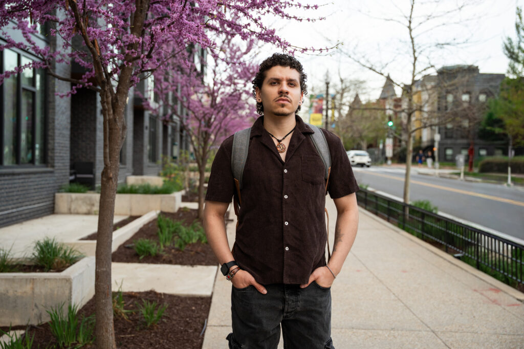 Yousef stands on the sidewalk looking at the camera with his hands in his pockets, backpack on.