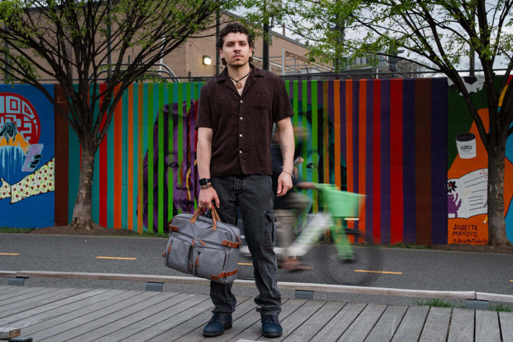 Yousef stands on a walkway looking at the camera, holding a duffle bag. A colorful mural is in the background as well as a cyclist riding by